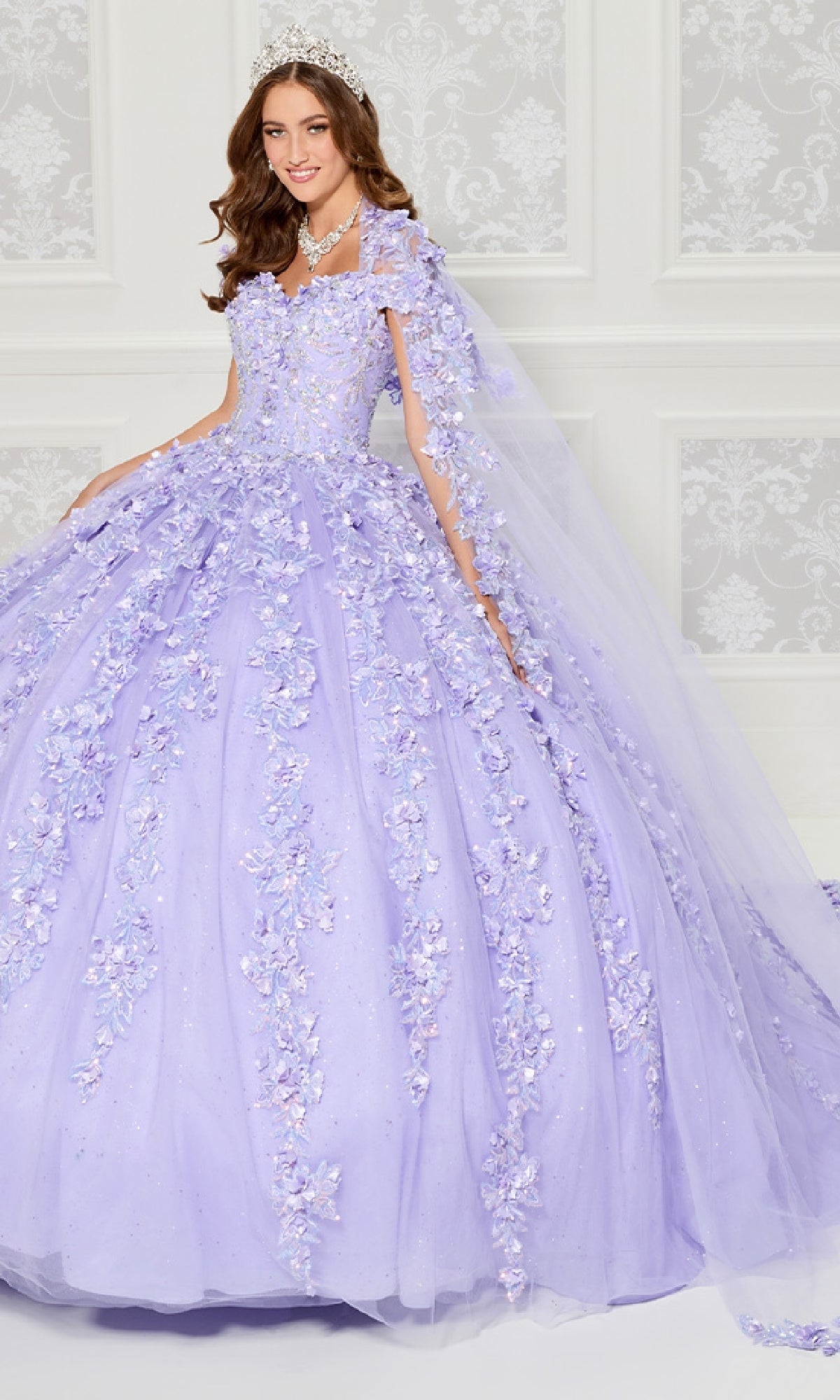A woman in the Princesa PR30120 lavender floral tulle quinceanera dress with 3D appliqués, tiara, and necklace stands before an ornate white wall.